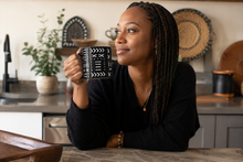 A woman with long black braids sits at a kitchen counter holding a black ceramic mug with white geometric patterns. She is wearing a black long-sleeve top and looking thoughtfully to the side.