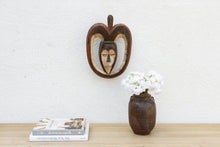 Decorative mask on a wall above a vase with flowers and books on a wooden surface.