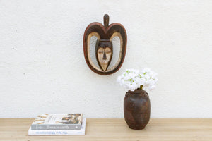 Decorative mask on a wall above a vase with flowers and books on a wooden surface.