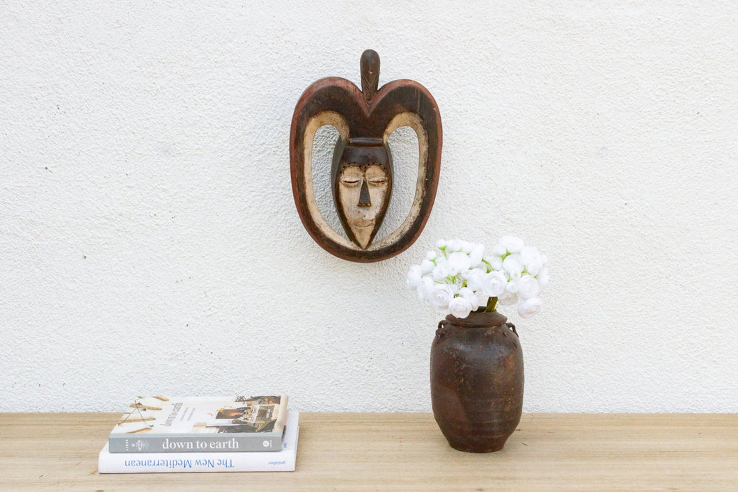 Decorative mask on a wall above a vase with flowers and books on a wooden surface.