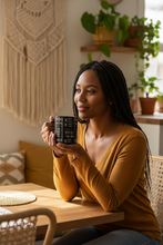 A woman with long black braids sits at a wooden table in a cozy room, holding a black mug with white text. She is wearing a mustard yellow long-sleeve top and blue jeans.