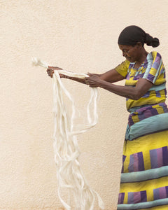 Woman in colorful traditional african attire holding sisal strands against a beige wall