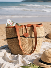 Woven beach bag with leather straps on a sandy beach with ocean waves in the background.