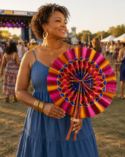 A woman in a blue dress holds a large, colorful folding fan with a vibrant geometric pattern in pink, orange, yellow, and blue. The background shows an outdoor festival scene with people and a stage at sunset.