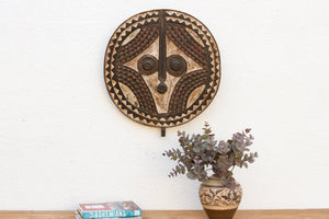 Decorative wall mask on a white wall with a vase of flowers and books below.