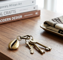 Brass cowrie shell keychain with keys on a wooden surface next to books and a bowl.