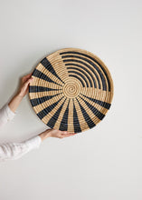 Person holding a woven round mat with black and beige stripes on a light gray background