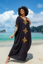 Woman in a black dress with gold embroidery standing on a beach with ocean and sky in the background