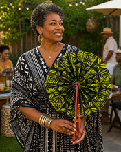 A woman outdoors holding a black and olive green patterned folding umbrella, wearing a black and white patterned dress and gold jewelry.