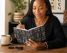 A woman with braided hair sits at a wooden table, smiling while writing in a black notebook with white geometric patterns. She wears a black long-sleeve top and gold hoop earrings.