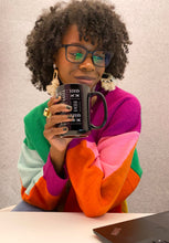 A woman with curly hair and glasses holds a black mug with white text, wearing a colorful color-blocked sweater in green, pink, orange, and light blue, against a neutral gray background.