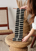 Person holding a black and white patterned african fabric wall art. A cork stool with a wooden chair and woven basket in the background.