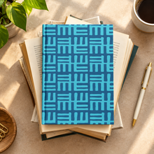 A hardcover notebook with a blue and turquoise geometric patterned cover, resting on a stack of open books on a beige desk with a plant, coffee, and a pen nearby.
