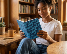 A woman with curly hair sits at a wooden table in a cozy library or cafe, reading a book with a blue geometric patterned cover. She is wearing a white t-shirt and blue jeans, with a small plant and a steaming mug on the table next to her.