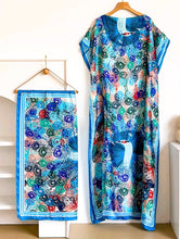 Colorful peacock-patterned kaftan dress and scarf on hangers against a white background