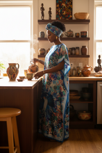Woman in a teal blue kaftan dress standing in a kitchen with various pots and vases.