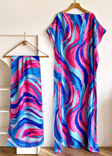 A matching two-piece set featuring a long kaftan dress and a coordinating scarf, both decorated with a vibrant abstract wavy pattern in pink, blue, and light blue. The dress is displayed on a wooden hanger against a neutral wall, while the matching scarf is draped over a white stand to the left.