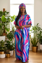 A woman stands indoors wearing a flowing kaftan dress and matching headwrap with a bold abstract swirl pattern in pink, blue, and white. The room has light wood floors and multiple potted green plants around her.