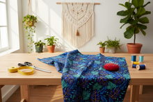 Table with blue fabric, sewing tools, and plants in a bright room.