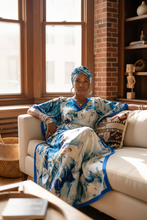 Woman in a blue and white kaftan dress sitting on a couch in a room with large windows and a brick wall.