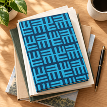 A stack of notebooks and books on a wooden desk, with a blue and turquoise geometric patterned notebook on top. A black and gold pen, a potted plant, and a ceramic cup of coffee are arranged around it.