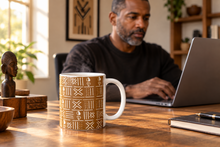 A white ceramic mug with a brown geometric tribal pattern sits on a wooden desk in a home office setting, with a man working on a laptop in the background.