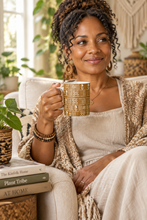 A woman sitting in a cozy living room setting, holding a patterned beige and brown ceramic mug. She is wearing a cream ribbed top and a textured beige cardigan.