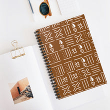 A brown spiral notebook with a white geometric patterned cover, styled on a white desk with an open book, sunglasses, a gold binder clip, and a pen.