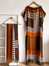 Houndstooth patterned scarf and kaftan dresswith brown and orange accents on a hanger against a white wall.