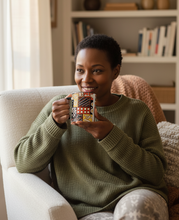A woman sitting in a cozy living room on a cream armchair, holding a patterned ceramic mug. She is wearing an olive green waffle-knit sweater and patterned leggings.