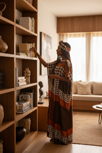 Woman in kaftan dress  reaching for a book on a wooden shelf in a well-lit room.