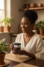 A smiling woman with curly hair sits at a wooden table in a warm, sunlit kitchen or cafe setting, holding a black and white patterned ceramic mug.