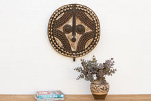 Decorative wall mask above a wooden shelf with books and a vase.