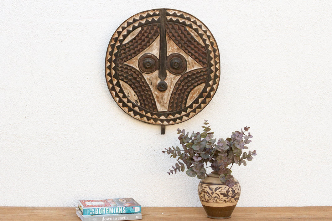 Decorative wall mask above a wooden shelf with books and a vase.