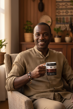 A smiling man wearing a beige button-up shirt and matching trousers sits in a woven armchair, holding a white mug with a black and red geometric pattern. The background shows a warm, cozy interior with wooden furniture and plants.