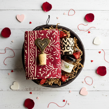 Gift basket with a red patterned towel, bottle, and decorative items on a white wooden surface with heart decorations.