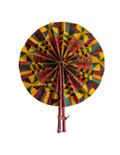 A round african fabric hand fan with a colorful geometric Kente pattern in yellow, green, red, and black, attached to a brown leather handle on a plain white background.