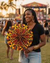 A woman at an outdoor festival holds a colorful fabric fan with a bold geometric pattern in yellow, green, red, and black. She is wearing a black t-shirt and light blue denim shorts, with a blurred crowd and stage in the background.