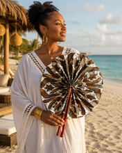 A woman on a sandy beach holds a brown and white patterned fabric fan. She is wearing a white crochet boho tunic, gold hoop earrings, and gold bangles, with the ocean and beach cabana in the background.