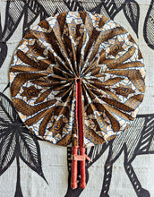 A circular fabric hand fan with a brown, gold, and white batik pattern, featuring a red handle and two red tassels, laid flat on a black and white patterned textile background.