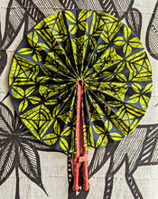 A folding hand fan with a vibrant lime green and black leaf pattern, featuring red plastic handles, displayed flat against a matching black and beige patterned fabric background.