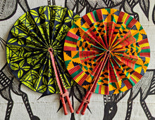 Two round fabric hand fans with leather handles, displayed flat against a black and white patterned background. The fan on the left features a black and lime green geometric pattern, and the fan on the right features a red, yellow, and green Kente-style pattern.