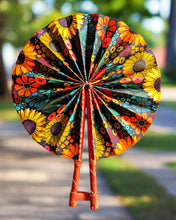 Colorful floral-patterned hand fan with wooden handle against a blurred outdoor background