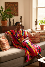 Woman in a colorful kaftan dress sitting on a couch in a living room with plants and decor.