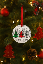 Circular ornament with tree designs hanging on a Christmas tree with red ribbon and other ornaments.