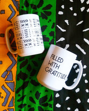 Two white ceramic mugs with black printed designs, one featuring geometric patterns and the other reading 'FILLED WITH GRATITUDE', displayed on a colorful patterned fabric background.