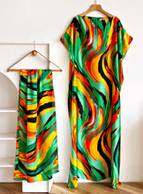 A matching short-sleeve kaftan dress and a coordinating scarf/belt both decorated with a vibrant, abstract wavy pattern in green, yellow, orange, red, and black. The dress is displayed on a wooden hanger against a white wall, while the skirt hangs on a wooden clothes rack.