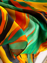 A close-up of a lightweight, semi-sheer fabric with an abstract, painterly pattern in vibrant green, orange, yellow, and black.