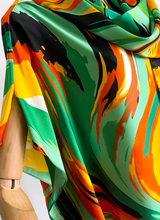 A close-up of a glossy silk scarf draped on a wooden mannequin, featuring a vibrant abstract print with swirls of green, orange, yellow, black, and white.