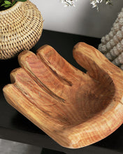 Wooden hand-shaped bowl on a dark surface with a woven basket and decorative items in the background.